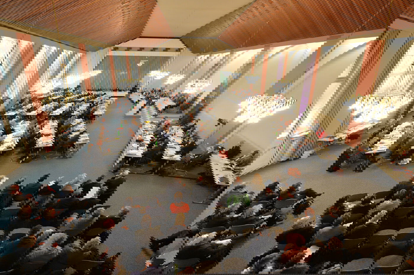 Ein großer Saal mit vielen Stuhl und Tischreihen, an denen Menschen sitzen. An der gegenüberliegenden langen Seite steht ein Redepult. 