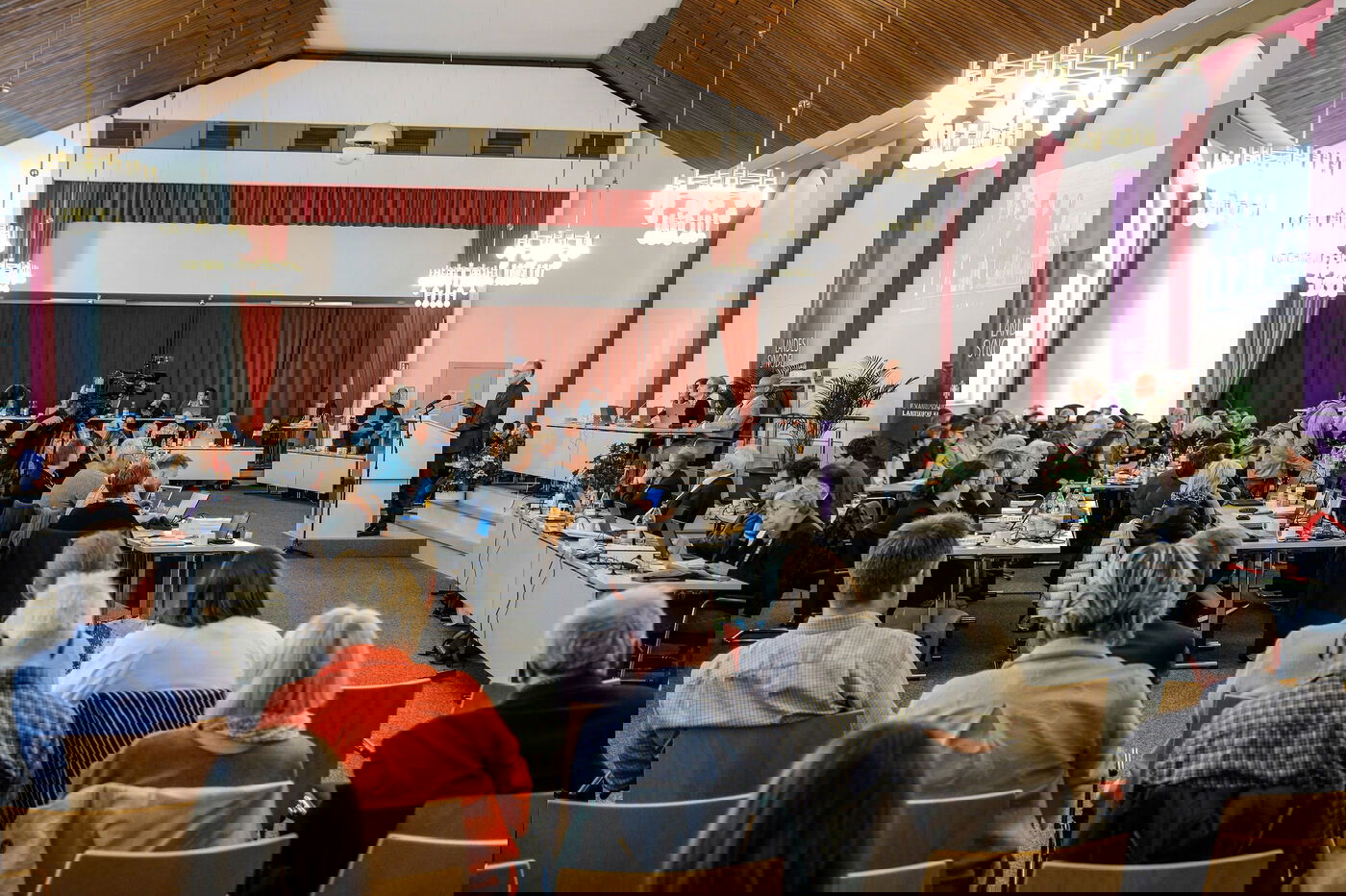 Ein großer Saal mit vielen Stühlen, auf denen Menschen in Reihen hintereinander sitzen. An einer der langen Seiten steht ein Podium mit einer männlich gelesenen Person dahinter. 