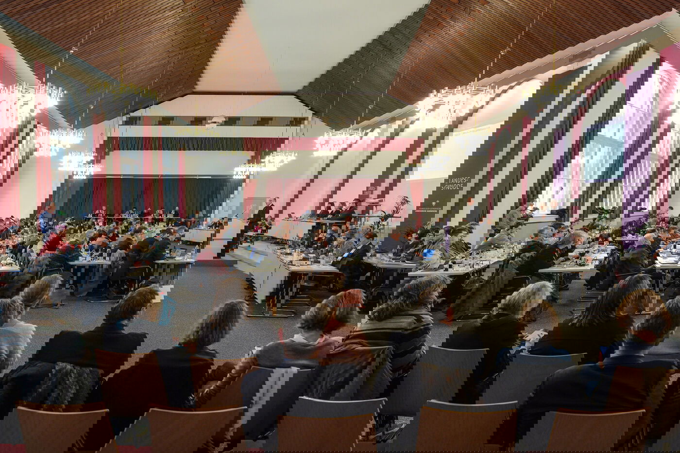 Ein großer Sitzungssaal mit vielen Menschen, die auf Stühlen sitzen. Am rechten Rand ist ein Podium.