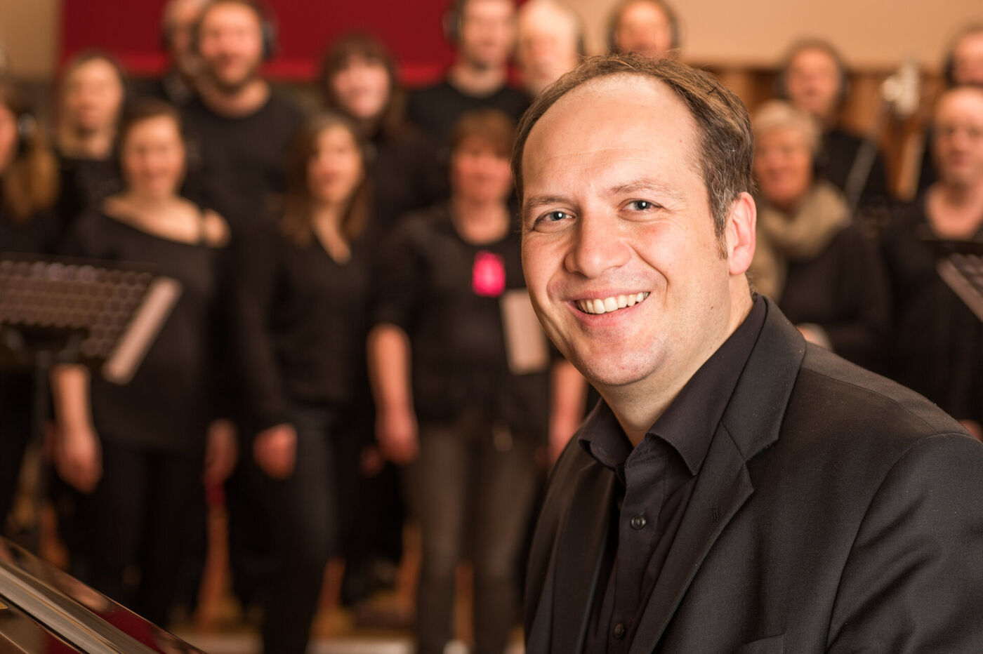 Eine als Mann lesbare Person im schwarzen Hemd steht im Vordergrund, im Hintergrund ist unscharf ein Chor zu sehen.