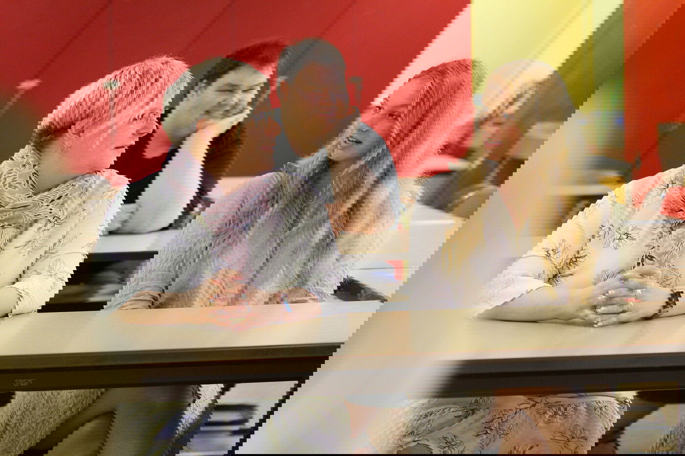 Eine Frau und eine junge Frau sitzen an einem Tisch vor einer roten Wand, hinter ihnen sitzt ein junger Mann.