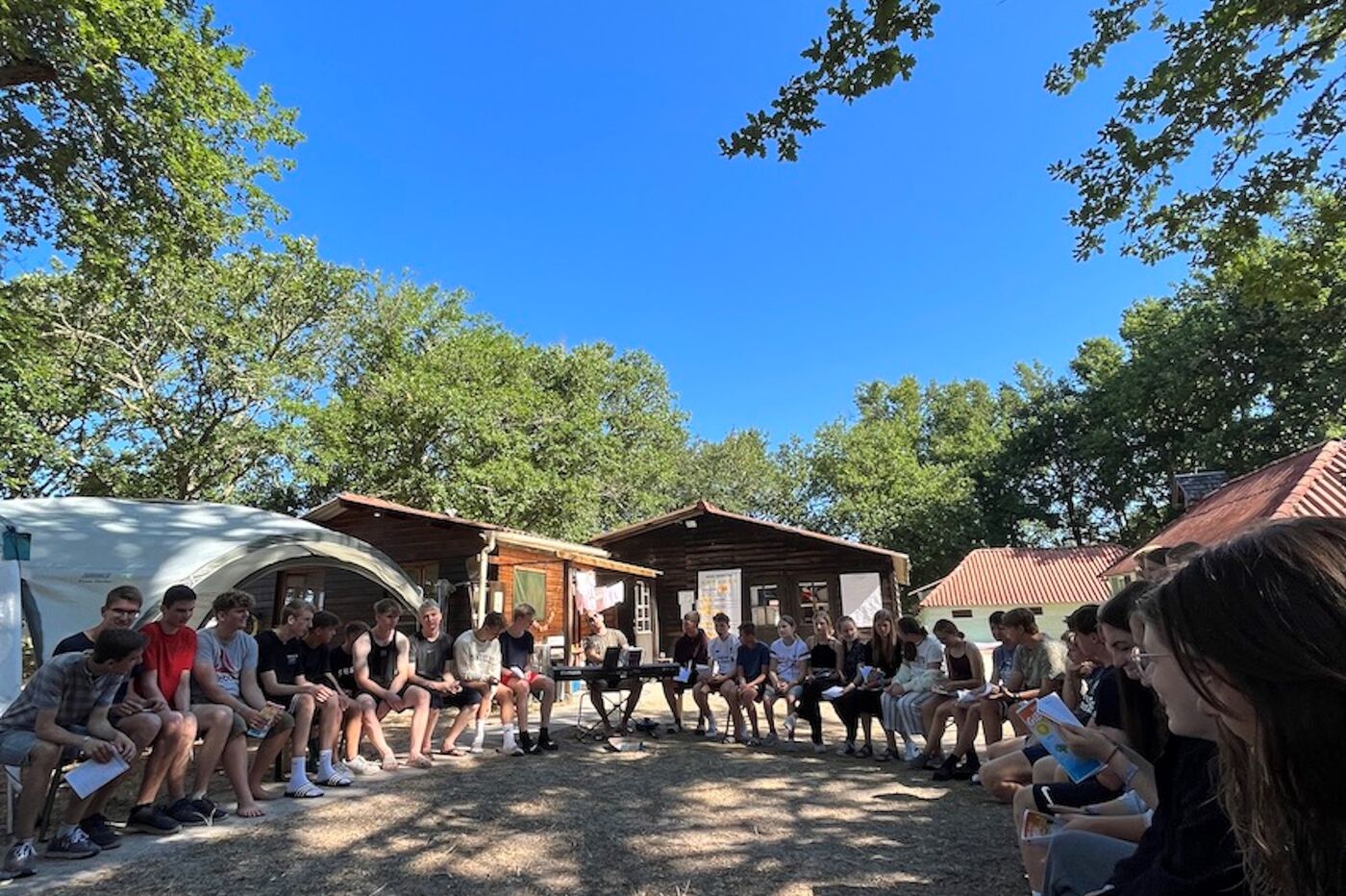 Unter strahlend blauem Himmel und grünen Bäumen sitzen junge Menschen in einem großen Kreis.