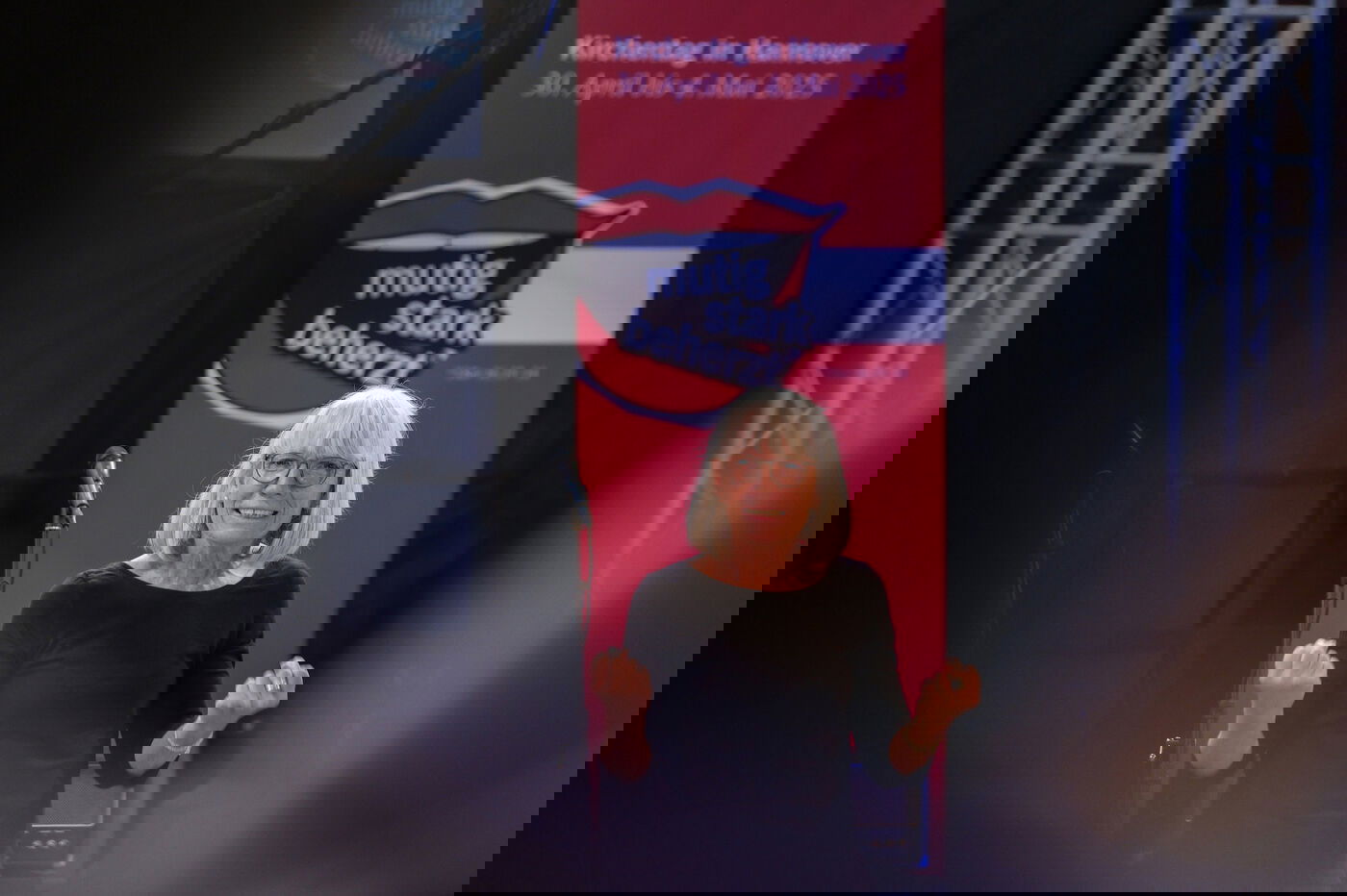 eine weiblich gelesene Person in schwarzem Pulli und mit blonden Haaren steht auf einer Bühne und gebährdet gesungenen Text.