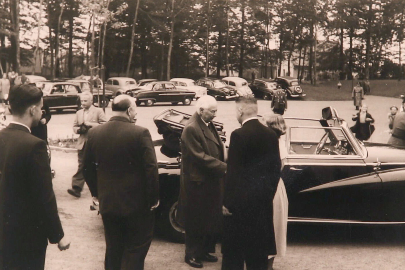 Historische Aufnahme mit mehreren Menschen vor einer Staatskarosse auf einem Parkplatz.