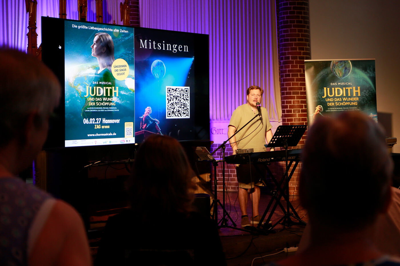 Eine als Mann lesbare Person steht am Keyboard und spricht in ein Mikrofon. Links neben ihm steht ein Rollup zum Chormusical „Judith“.