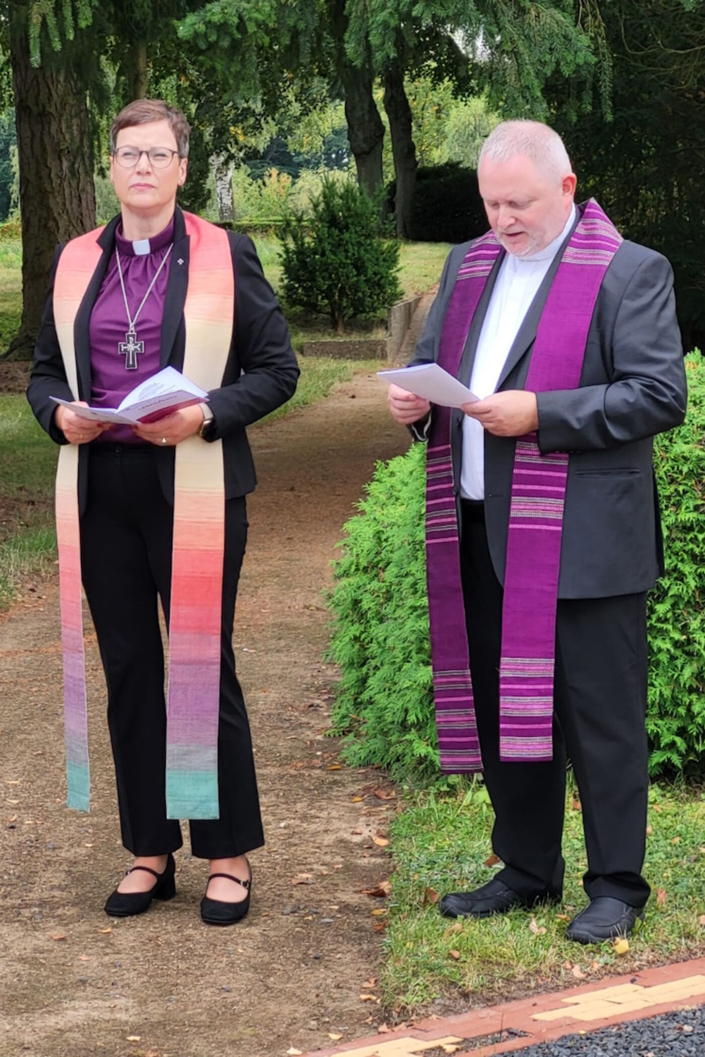 Eine als Frau und eine als Mann lesbare Person im Talar stehen auf einem Friedhof und sprechen ein Gebet.