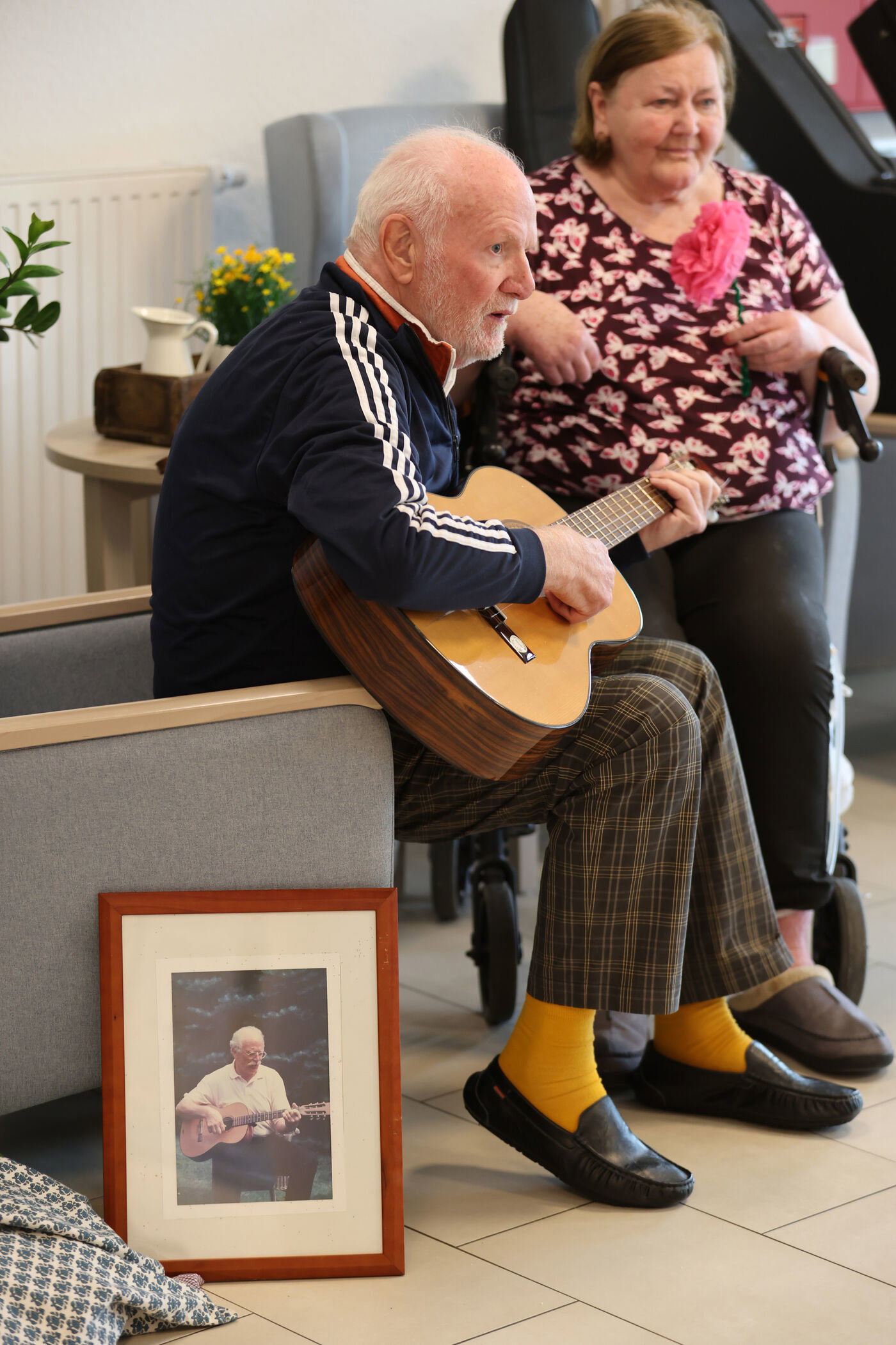 Eine als Mann lesbare ältere Person spielt in einem Sessel Gitarre. An dem Sessel lehnt ein gerahmtes Foto von ihm mit Gitarre.