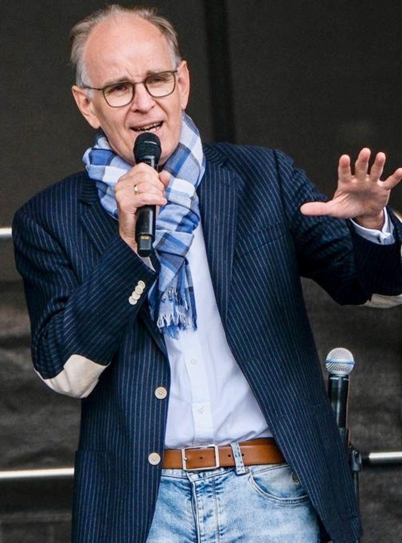 Das Bild zeigt eine männlich lesbare Person ohne Bart, mit kurzen grauen Haaren, einer Brille mit dunkler Fassung, einem hellblauen Schal, weißem Hemd und dunklem Sakko. Die Person hält ein Mikrophon in ihrer rechten Hand und hält die linke Hand auf Brusthöhe.
