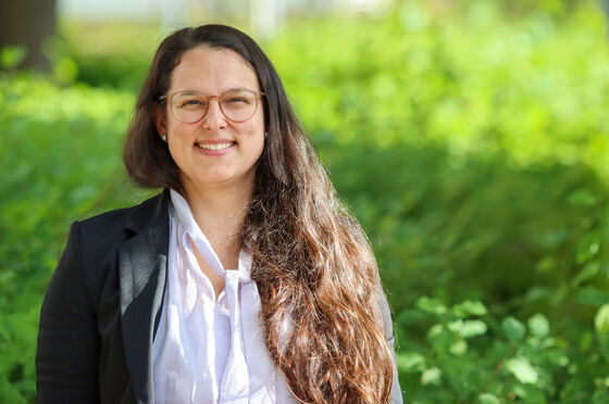 Eine Frau mit randloser Brille und langen, braunen Haaren trägt eine weiße Bluse und einen dunklen Blazer. Sie steht im Grünen und lächelt.