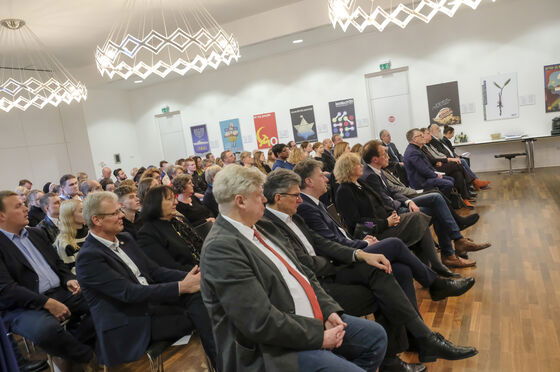 Mehrere dutzend Menschen sitzen in Stuhlreihen in einem großen Raum, viele tragen Anzüge und gute Kleidung. 