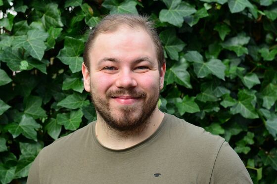 Eine männlich gelesene Person mit kurzem Bart und olivgrünem Shirt steht vor einer grünen Hecke.