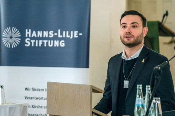 Eine männlich gelesene Person mit dunklem Pulli und Jackett an einem Redepult. Im Hintergrund ein blau-weißer Aufsteller mit dem Schriftzug "Hanns-Lilje-Stiftung".