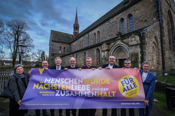 Eine weiblich zu lesende Person und sieben männlich zu lesende Personen halten ein Banner. Im Hintergrund ist eine Kirche zu sehen.