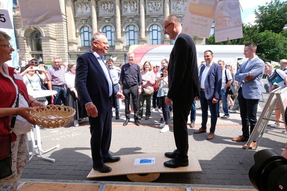 Zwei männlich lesbare Personen im Anzug stehen auf einer kleinen Wippe in Form eines Brettes, im Hintergrund stehen mehrere Menschen und schauen zu.