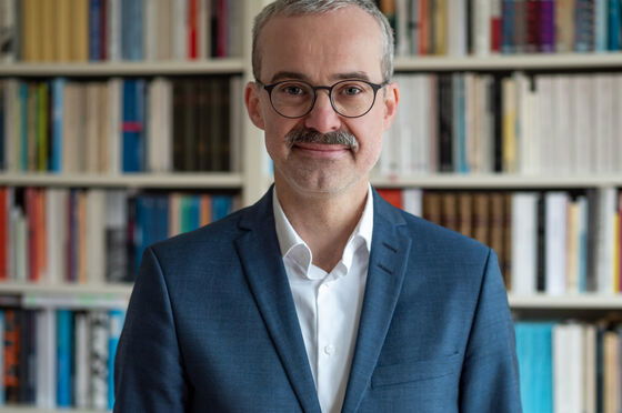 Das Bild zeigt eine männlich lesbare Person mit blauem Sakko, weißem Hemd, kurzen dunklen Haaren, Schnurrbart und Brille. 