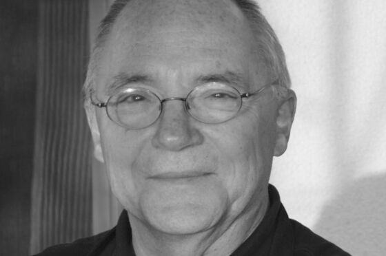 Eine männlich lesbare Person ohne Bart, mit Brille und kurzen grauen Haaren in einem dunklen Oberteil (sw-Bild).