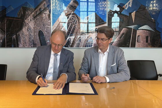 Zwei männlich zu lesende Personen sitzen an einem Tisch und eine Person unterschreibt ein Dokument, das auf dem Tisch liegt.