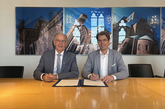 Zwei männlich zu lesende Personen sitzen an einem Tisch und vor ihnen liegt ein Dokument zur Unterschrift.