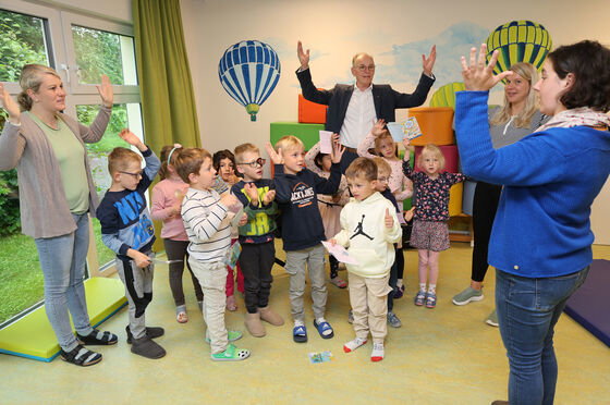Eine Gruppe Kinder singt gemeinsam mit einer als Frau und einer als Mann lesbaren Person. Alle heben die Hände in die Höhe.