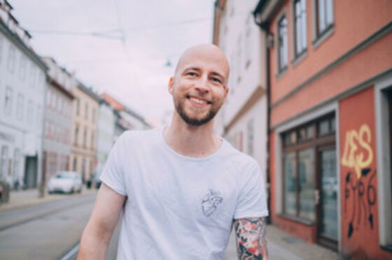 Eine männlich gelesene Person steht im weißen T-Shirt draußen auf der Straße; er hat einen Vollbart und eine Glatze; er lacht.