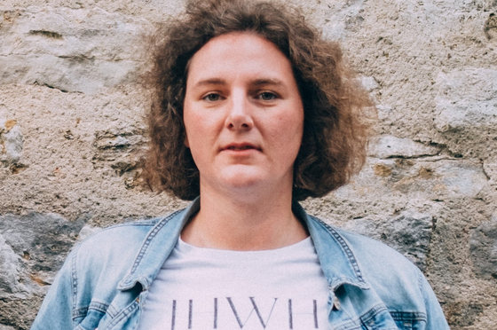 Eine weiblich gelesene Person steht vor einer Mauer. Sie hat gelockte, schulterlange braune Haare. 