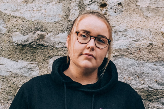 Eine weiblich gelesene Person steht draußen vor einer Mauer; sie hat ihre Haare zurückgebunden und trägt eine Brille. 