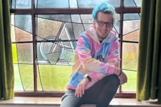 Eine männlich gelesene Person sitzt auf einer Fensterbank vor einem Kirchenfenster. Er hat bunte Kleidung hat und bunt gefärbte Haare. 