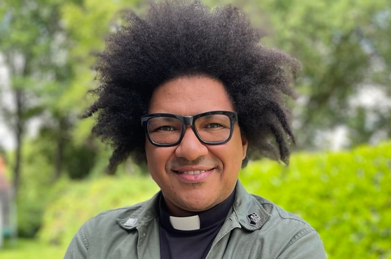 Eine männlich gelesene, schwarze Person mit dunklen gelockten Haaren und einer Brille mit dunkler Fassung. Er steht draußen. 