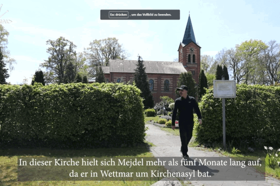 Ein Kiesweg führt durch eine grüne Hecke hindurch. Ein Mann mit Käppi läuft den Weg entlang, im Hintergrund ragt eine Kirche auf.