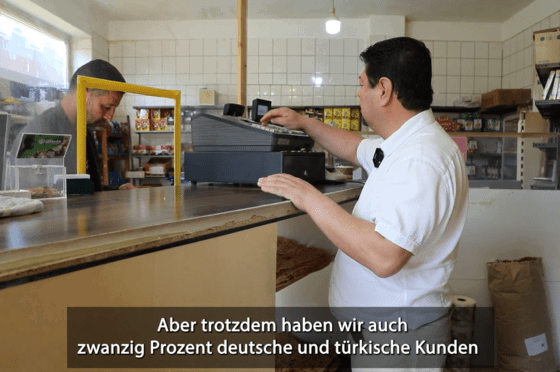 Ein Mann in weißem Shirt steht an einer Theke. Auf der anderen Seite steht ein anderer Mann in braunem Hemd. 