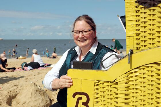 Eine Frau steht angelehnt an einen gelben Strandkorb. Sie hat eine blaue Weste an und eine weisse Bluse. Sie hat lange, dunkelblonde Haare und eine Brille. Im Hintergrund sieht man das Meer und paar Menschen am Strand.