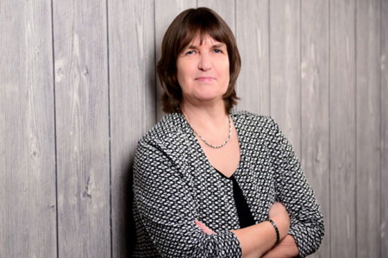 Eine weiblich gelesene Person mit schwarz-weißer Bluse und kurzen braunen Haaren lehnt an einer hölzernen Wand.