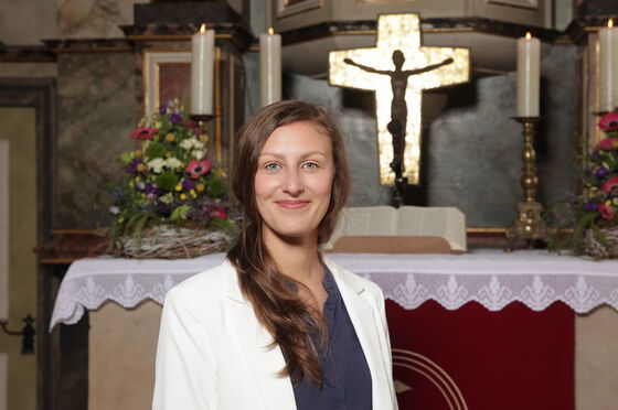 Eine junge Frau mit weißer Jacke und langen Haaren steht vor einem Altar mit goldenem Kreuz.