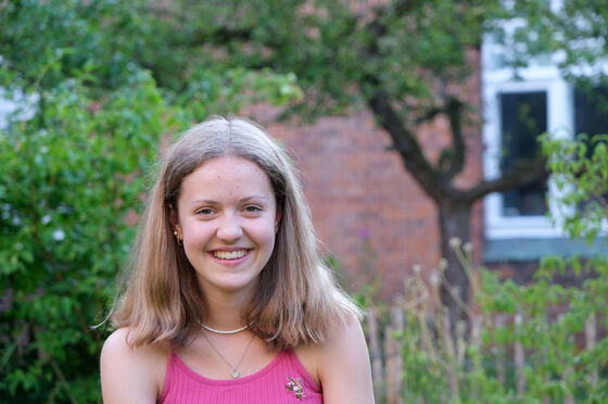 Eine junge Frau mit schulterlangen blonden Haaren und rosa Shirt lächelt, vor einem Backsteinhaus stehend, vor dem ein Baum wächst.