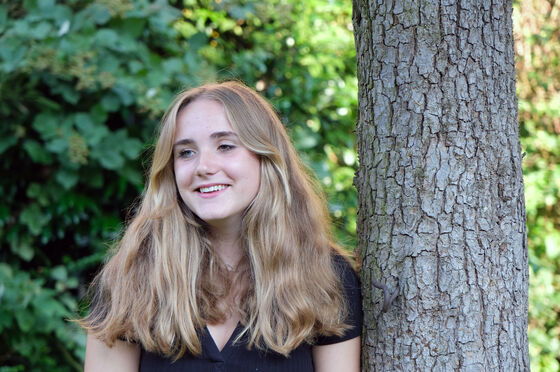 Eine junge Frau mit langen blonden Haaren lehnt an einem Baumstamm.