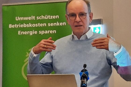 Ein Mann mit Halbglatze und Brille in grauem Pulli über einem Hemd steht an einem Redepult und gestikuliert.