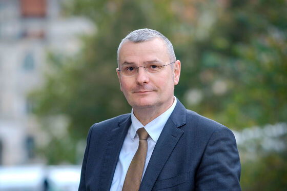 Eine männlich gelesene Person in blauem Jackett und mit Krawatte, mit raspelkurzen grauen Haaren und Brille, guckt aufmerksam in die Kamera.