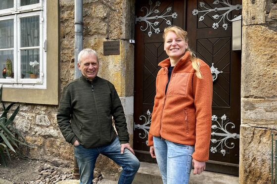 Eine weiblich gelesene Person mit orangefarbener Jacke und langen blonden Haaren steht mit einer männlich gelesenen Person in Outdoor-Jacke vor einer Haustür mit dem Schild "Kloster Loccum Forstverwaltung".