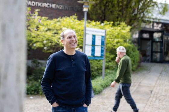 Eine männlich gelesene Person in dunklem Shirt steht draußen und lacht.