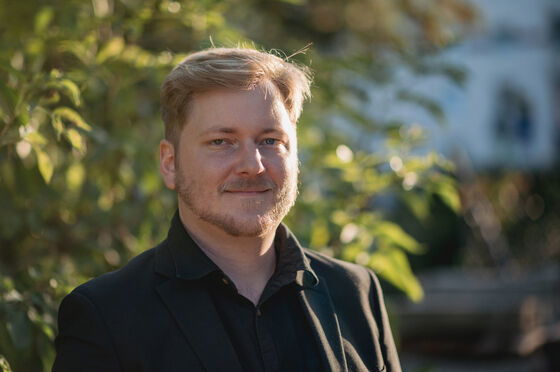 Eine männlich gelesene Person mit kurzen blonden Haaren in dunklem Anzug steht vor einem grünen Strauch.