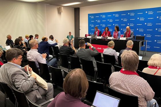 Menschen sitzen in Stuhlreihen und schauen auf ein Plenum, auf dem vier Menschen sitzen.