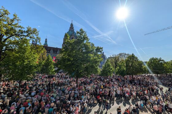 Tausende Menschen stehen und sitzen im Sonnenschein auf einem asphaltierten Platz mit vielen Bäumen drumherum.