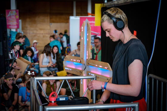 Eine Person mit Kopfhörern steht an einem DJ-Pult, andere Personen schauen ihr zu.