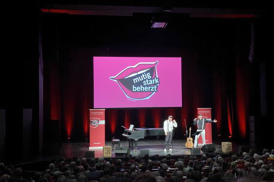 Auf einer Bühne, über der das Logo des Kirchentags eingeblendet ist, stehen ein Flügel und zwei männlich zu lesende Personen.