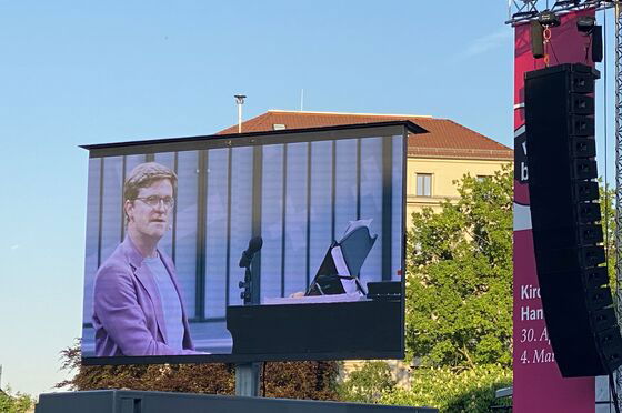 Eine männlich zu lesende Person, die an einem Flügel sitzt, ist auf einer Großleinwand zu sehen.