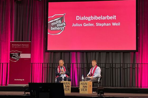 Zwei männlich zu lesende Personen sitzen auf einer Bühne und sprechen in Mikrofone.