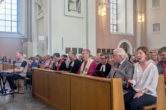Menschen sitzen in Kirchenbänken in einer Kirche. In der ersten Reihe sitzen mehrere Personen, die kirchliche Gewänder tragen.