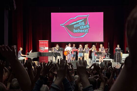 Eine dunkle Bühne, darauf im Licht eine Band mit Jugendlichen; im Hintergrund das Logo des Kirchentages mit einem Kussmund und den Worten "Mutig, Stark, Beherzt"