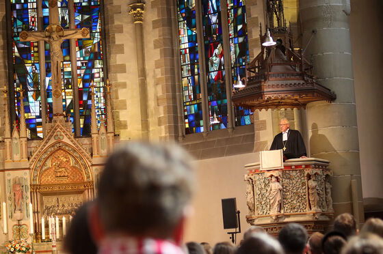 Eine männlich gelesene Person auf einer Kanzel, im Hintergrund bunte Kirchenfenster. Davor viele Menschen von hinten zu sehen.
