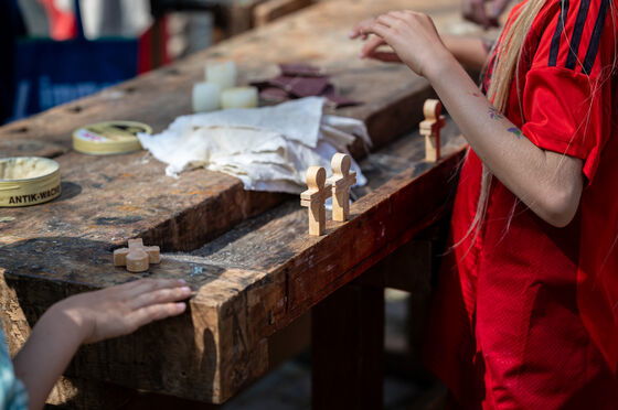 Auf einer Werkbank stehen mehrere kleine Holzkreuze, daneben erkennbar ein jüngerer Mensch, der an den Kreuzen schleift. 